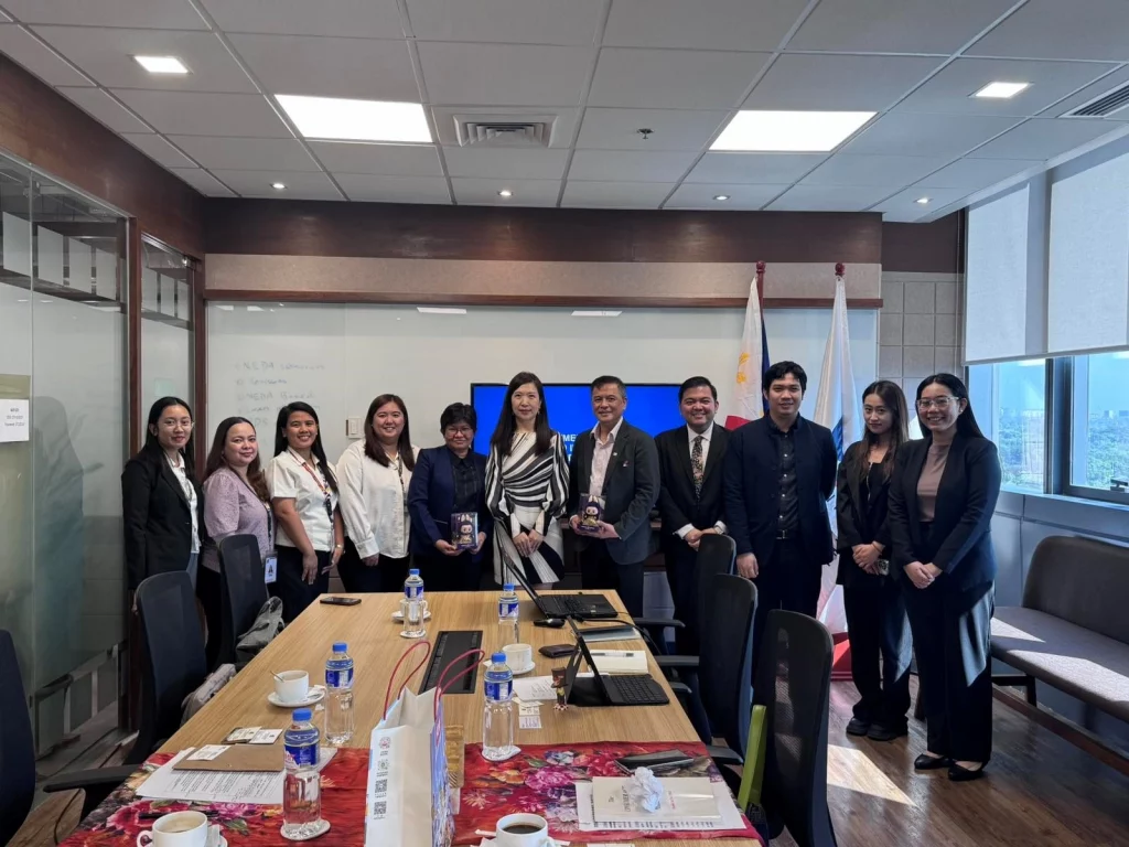 Individuals are standing behind a long conference table in a meeting room with large windows showing a cityscape. Two Philippine flags are displayed behind them, along with another flag that is partially obscured. The individuals, identified in the caption as representatives from DTI-FTSC, BOI-IAS, CPAB, and officials from POP MART International Group, are dressed in business attire. Some are holding small, wrapped items. The setting suggests a formal meeting or collaboration. Given the context of POP MART's expansion in the Philippines and the presence of Philippine government agencies, it's likely that some individuals in the image are Filipino and others may be of Chinese descent, representing POP MART.