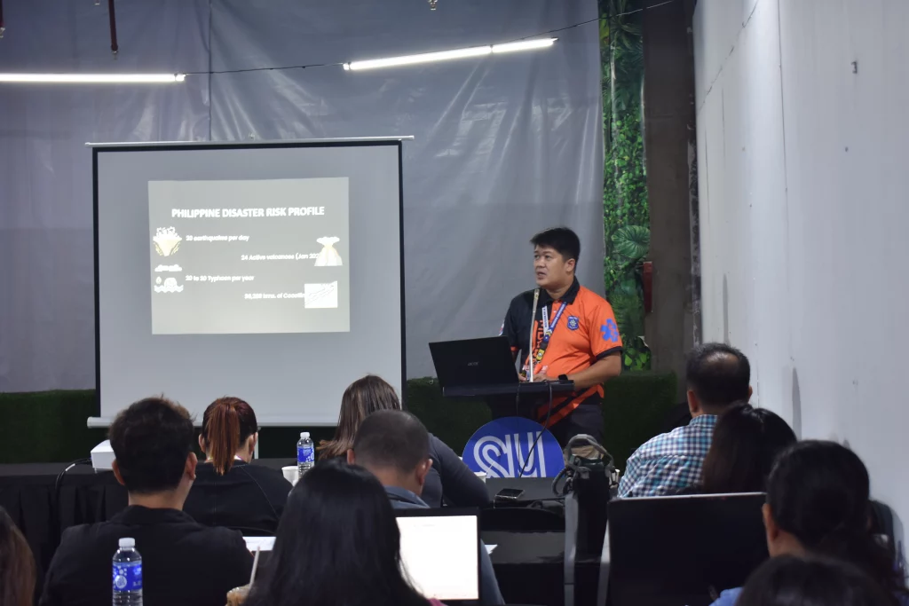 A man in an orange shirt is presenting on stage about the Philippine Disaster Risk Profile. A projector screen behind him displays the title. The SM logo is visible in the lower right corner.