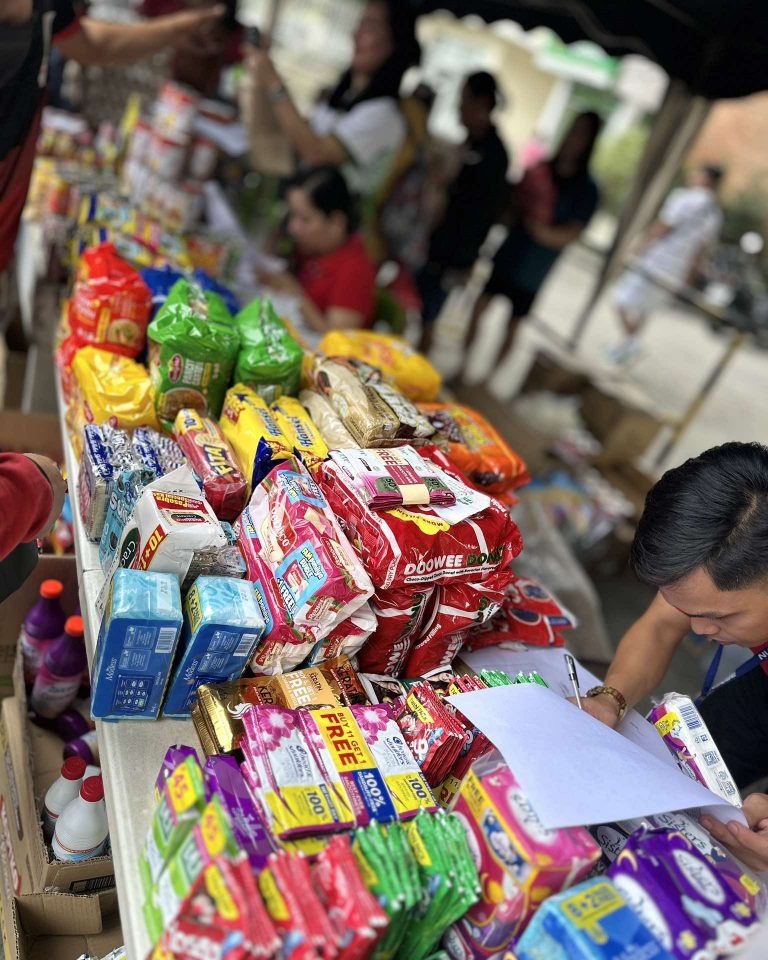 A close-up view of a table covered with an assortment of packaged goods, including snacks, cleaning supplies, and personal care items. Individuals are seen in the out-of-focus background.