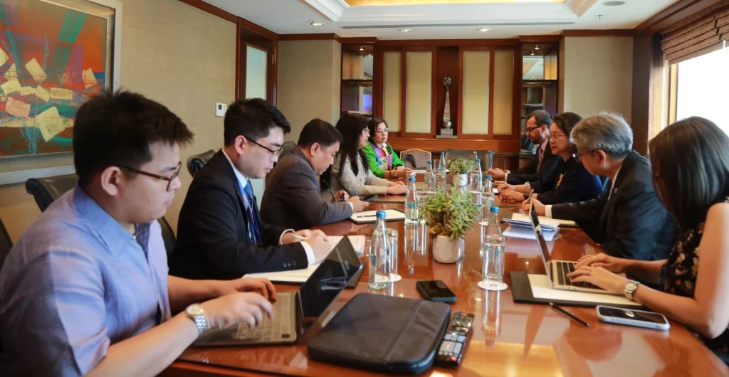A group of individuals are seated around a long wooden conference table in a well-lit room. Most of them appear to be working on laptops or looking at documents. The people seem to be engaged in a meeting or discussion. The photo caption indicates that these are representatives from the DTI (Department of Trade and Industry) and UNESCAP (United Nations Economic and Social Commission for Asia and the Pacific).