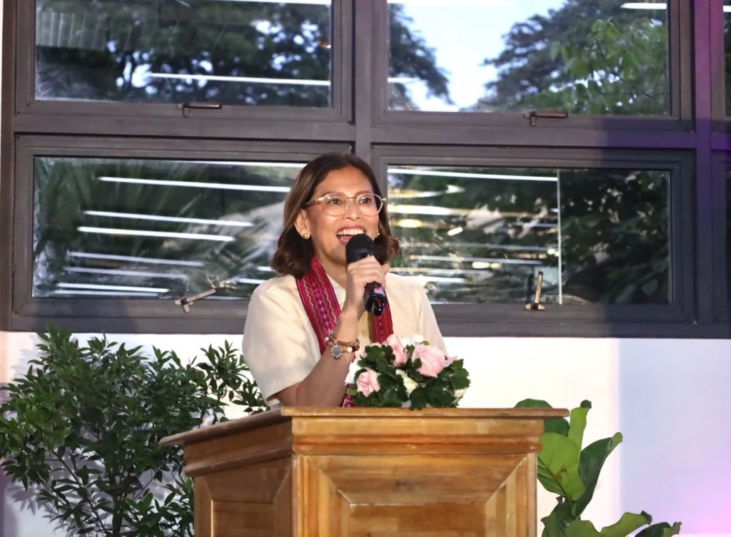 Cong. Stella Luz Quimbo stands behind a wooden podium, holding a microphone and speaking. The background includes large windows with greenery visible outside and some indoor plants near the podium.