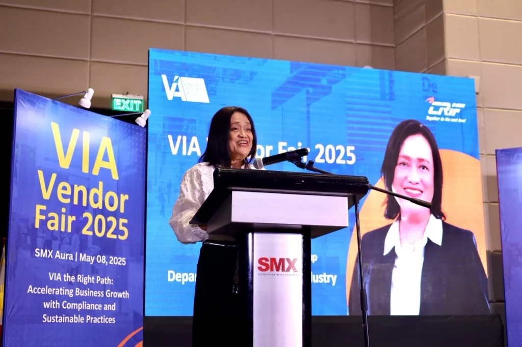 DTI Undersecretary Blesila Lantayona delivers a keynote speech at the podium during the VIA Vendor Fair 2025 at SMX Aura, with event banners and screens displaying the theme and date.