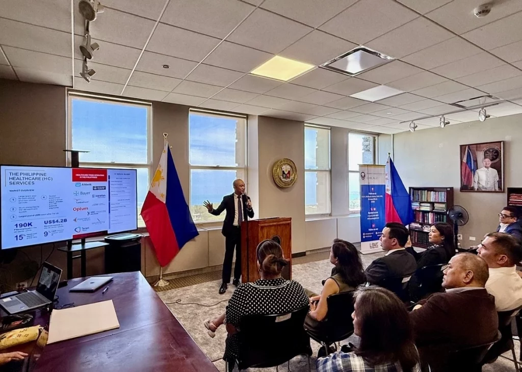 Mr. John Duenas presenting on the Philippine Healthcare Information Management Services at a seminar in Chicago, with a screen behind him displaying statistics on the Philippine startup ecosystem and two flags, including the Philippine flag, in the background.