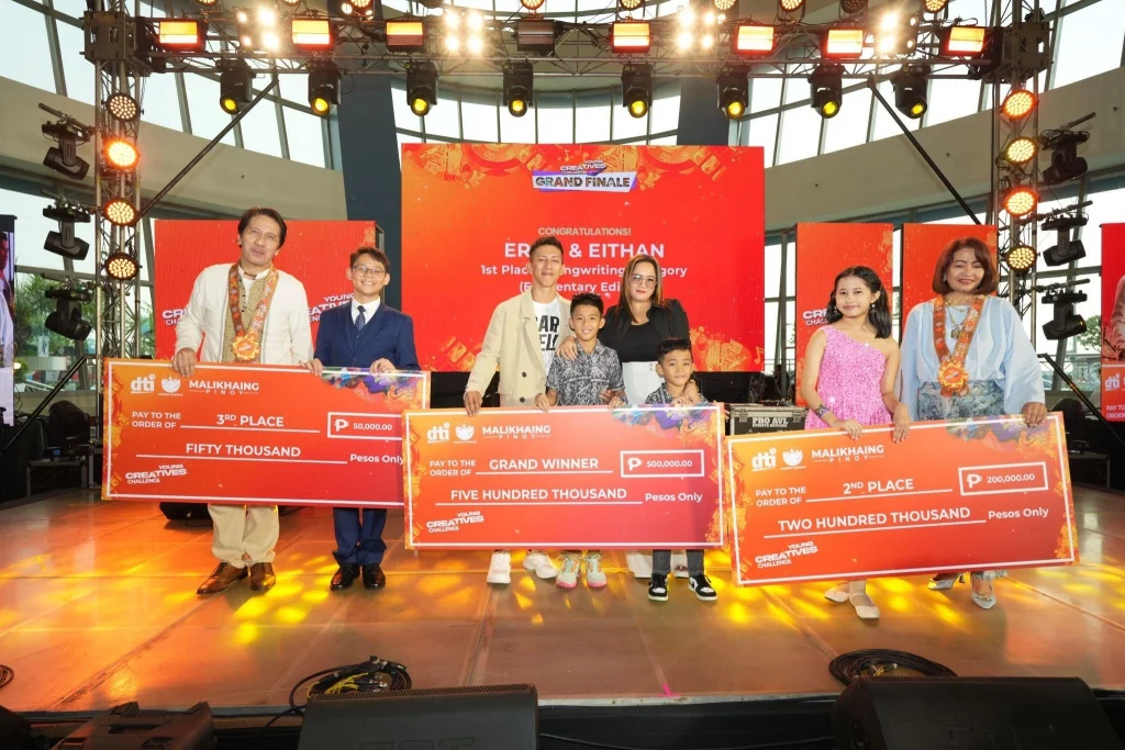 A group of individuals standing on stage holding large ceremonial checks during the Young Creatives Challenge Grand Finale. The backdrop displays a congratulatory message for "Earnest & Eithan" as 1st Place winners in the Songwriting Category. The checks show various prize amounts, including 50,000, 200,000, and 500,000 pesos, representing 3rd place, 2nd place, and Grand Winner positions.