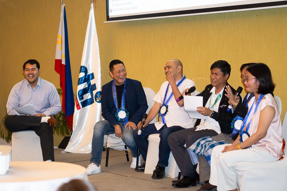 Panel speakers wearing formal attire and ceremonial ribbons during the Palawan Investors' Roadshow, seated on stage with a backdrop featuring the NIP flag and a presentation screen, highlighting discussions on energy efficiency for MSMEs.