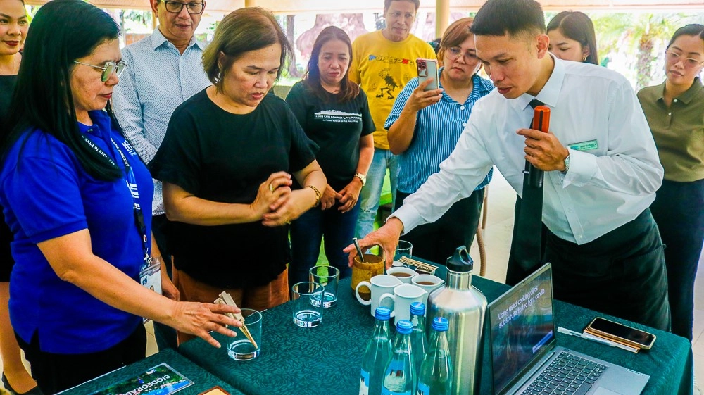 A group of people gathered around a table with bottles of water, a metal thermos, glass cups, and a laptop. One person is pouring liquid into a glass cup while others observe.