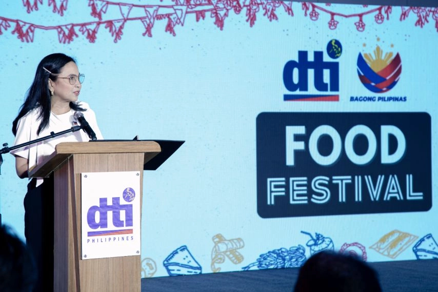 DTI Secretary Ma. Cristina A. Roque speaks at the podium during Food Festival 2025, with a backdrop displaying the DTI and Bagong Pilipinas logos, and illustrations of Filipino food items.