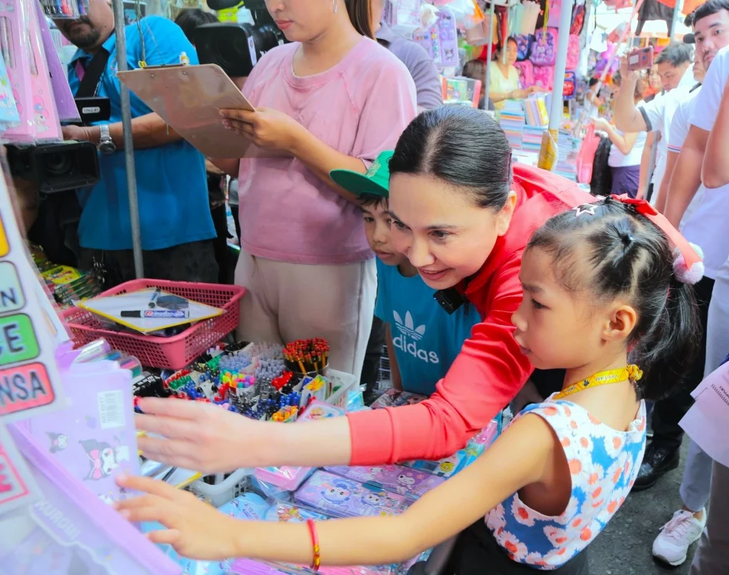 DTI Secretary Cristina A. Roque, accompanied by a child, inspects a school supplies stall in Divisoria Market, selecting from a variety of colorful notebooks, pens, and other essentials as part of the department’s price monitoring efforts.