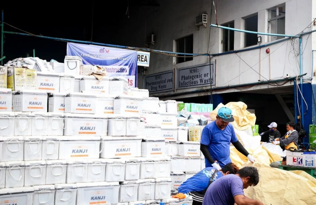 Workers in protective gear handle and organize stacks of confiscated automotive batteries labeled "KANJI" at a waste disposal facility in Valenzuela City. The activity is part of DTI’s ceremonial destruction of Php 110.7M worth of uncertified lead-acid batteries to ensure consumer safety and environmental compliance.