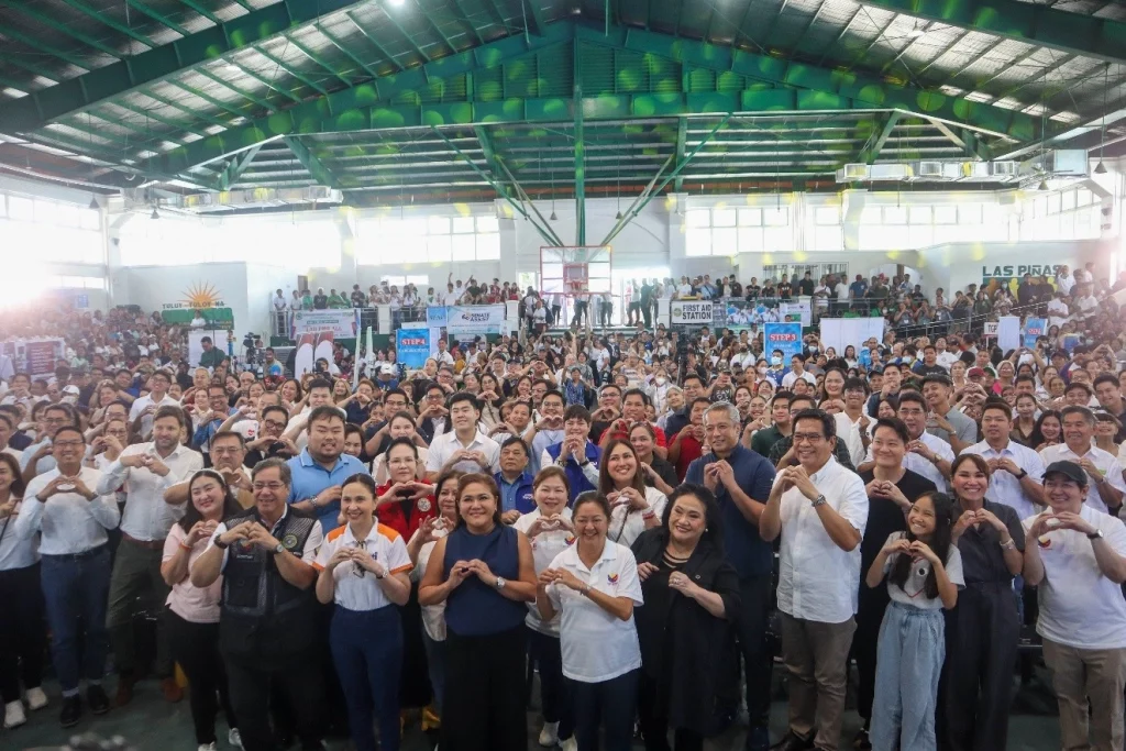A large crowd gathers at the 'Lab For All' event in Las Piñas, held in a spacious indoor venue with green beams and natural lighting. Attendees, including DTI representatives and community members, form heart shapes with their hands, celebrating the launch of the Diskwento Caravan and other public service initiatives.