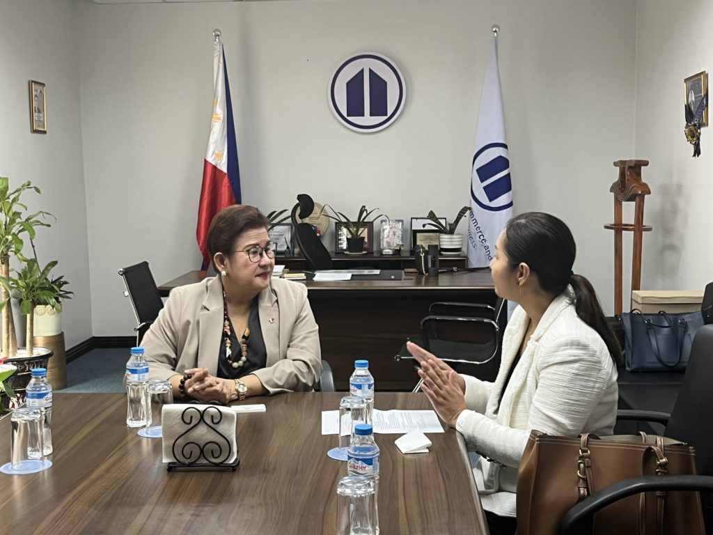 DTI Foreign Trade Service Corps Assistant Secretary Alma Argayoso (right), wearing a white blazer, sits at a conference table conversing with PCCI President Consul Eunina Mangio (left), who is wearing a brown blazer. The Philippine flag and PCCI logo are visible in the office background.