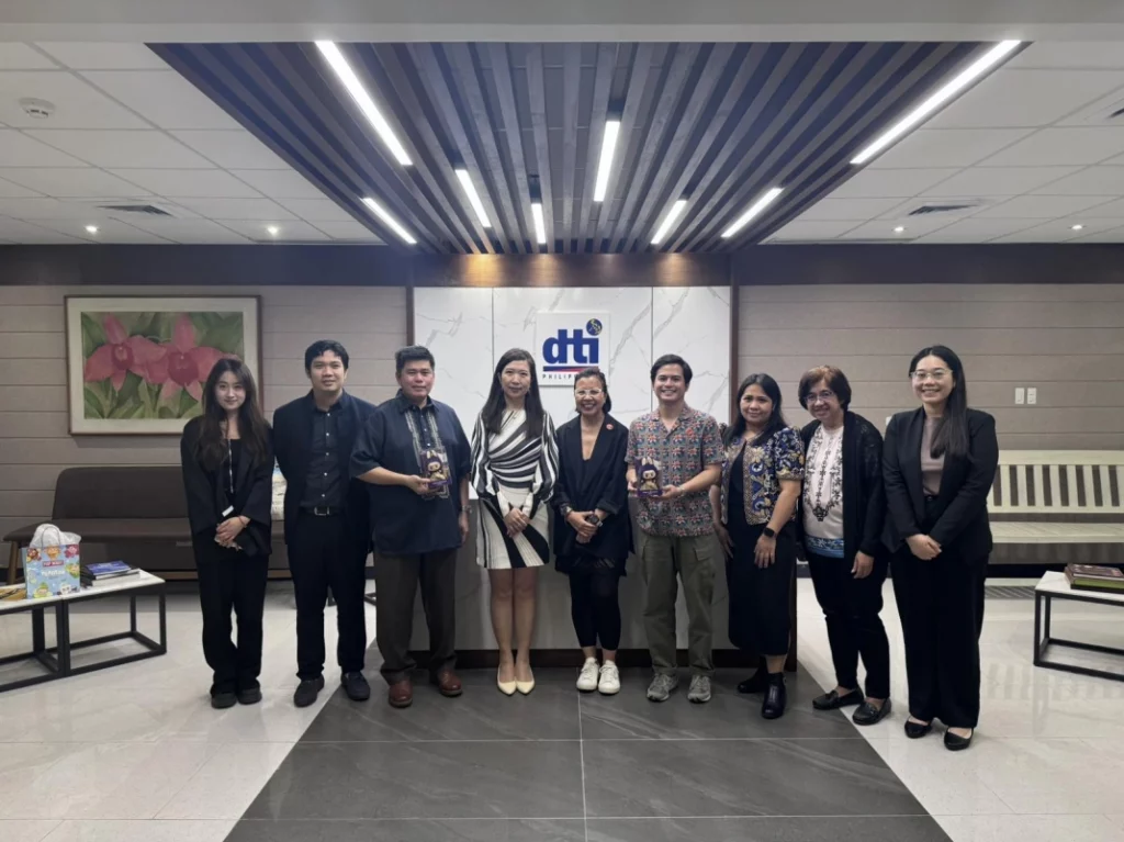 Individuals are standing behind a long conference table in a meeting room. The DTI logo is visible in the background. The individuals, identified in the caption as representatives from DTI-FTSC, DCP, IPOPHL, and officials from POP MART International Group, are dressed in business attire. Some are holding small, wrapped items. The setting suggests a formal meeting or collaboration. Given the context of POP MART's expansion in the Philippines and the presence of Philippine government agencies, it's likely that some individuals in the image are Filipino and others may be of Chinese descent, representing POP MART.