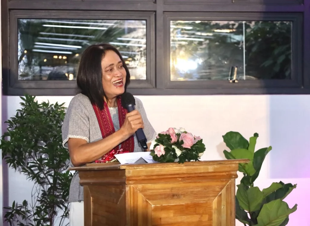Undersecretary Blesila Lantayon is standing behind a wooden podium, holding a microphone in her right hand and speaking. The podium has a small bouquet of pink flowers on it. There are plants in the background and a window with reflections of lights above her.