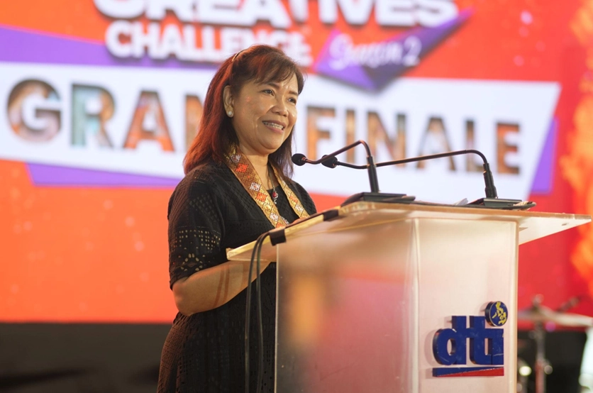 DTI Competitiveness and Innovation Group Assistant Secretary Leonila T. Baluyut delivering the opening message at the Young Creatives Challenge Season 2 Grand Finale, standing at a podium with the DTI logo and a colorful event banner in the background.