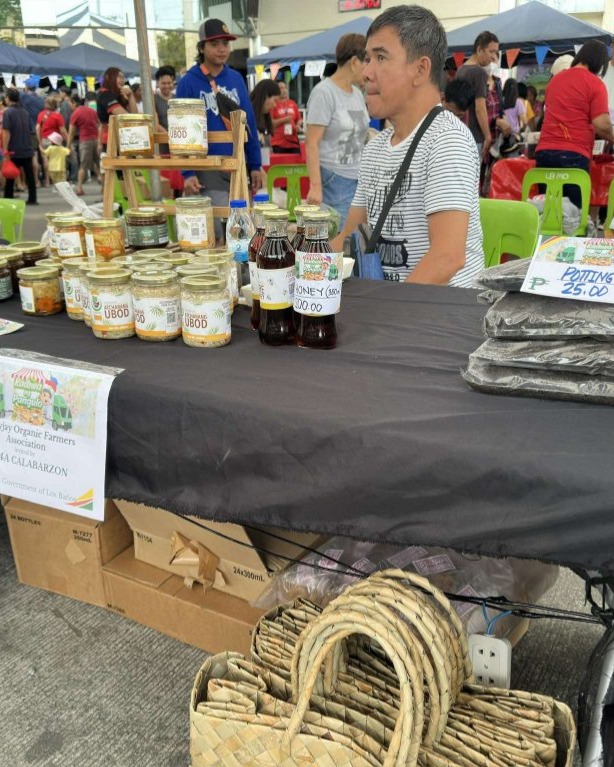 Local products from the Majayjay Organic Farmers Association CALABARZON are being sold at an outdoor market. The display includes jars of food, bottled drinks, and woven baskets. People are shopping in the background, indicating a community market or fair.