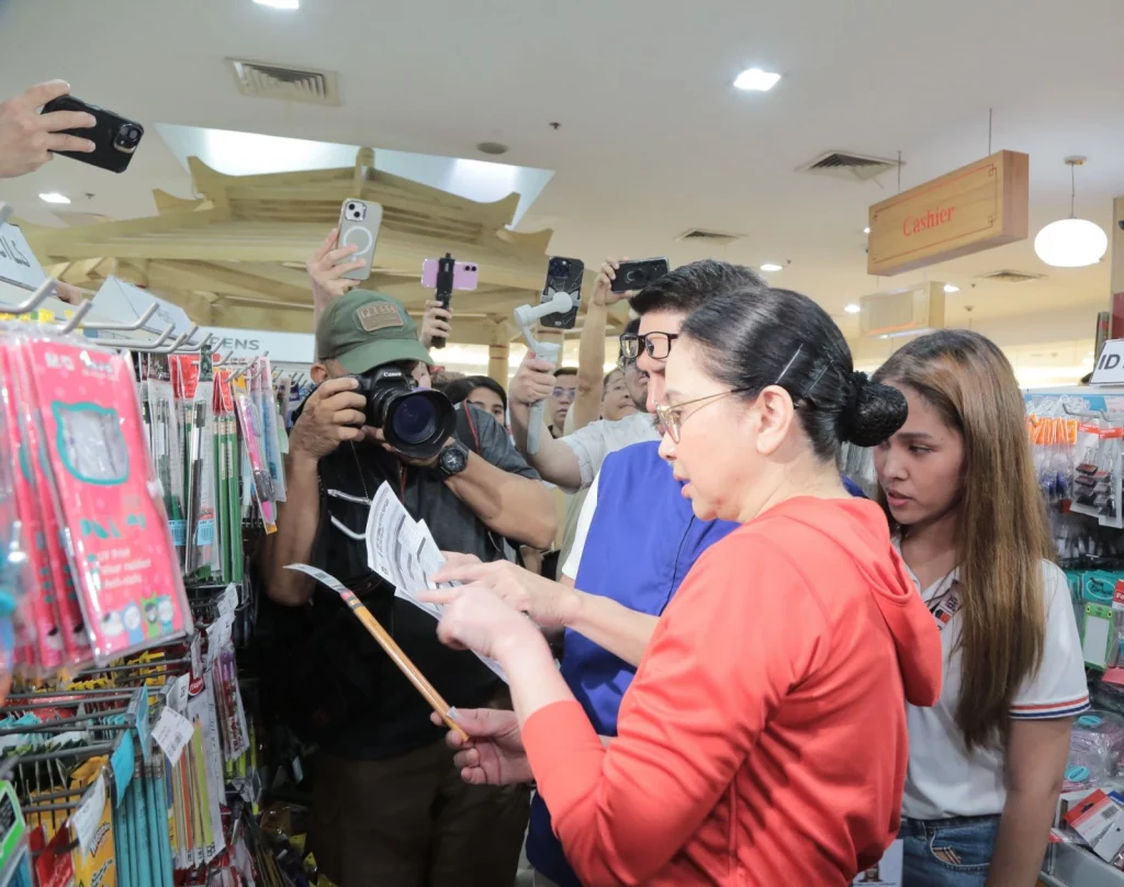 DTI Secretary Cristina A. Roque, holding a price guide, speaks with a store representative in a well-lit school supplies store, as onlookers document the inspection with their phones. Shelves are stocked with colorful stationery items under a 'Cashier' sign.