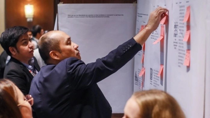 A group of individuals in formal attire is participating in a workshop or seminar. One person is placing or adjusting sticky notes on a board with several sheets of paper and additional sticky notes. The setting is professional, indicating a collaborative and interactive session.