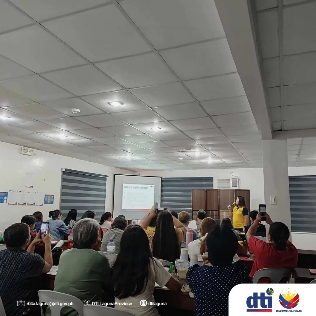 Participants attend a DTI-CMDI livelihood training session in Bay, Laguna, focusing on business compliance, licensing, and workplace efficiency. A speaker in a yellow top presents a slide while attendees take notes and photos.