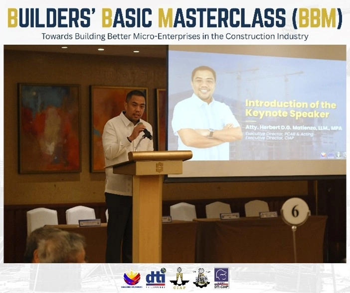 A man presenting at the Builders' Basic Masterclass (BBM) launch event, with a screen behind him displaying 'Introduction of the Keynote Speaker' and details about Atty. Herbert D.B. Matienzo, LL.M., MPA, Executive Director of PCAB and Acting Executive Director of CIAP. The venue is arranged with tables and chairs for attendees, including a table marked with the number 6.