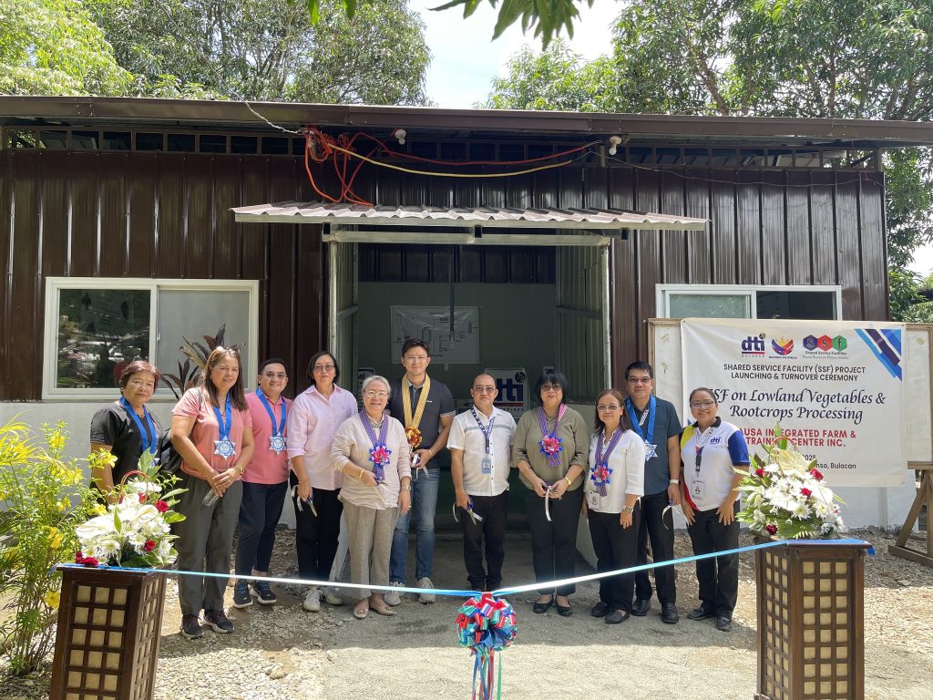 A group of people stands in front of a building during a ribbon-cutting ceremony for the Shared Service Facility (SSF) Project Launching & Turnover Ceremony on lowland vegetables and root crops processing at USAKA Integrated Farm & Training Center Inc. The building features dark brown walls and white-framed windows, with a banner detailing the event and decorative plants arranged around the area.