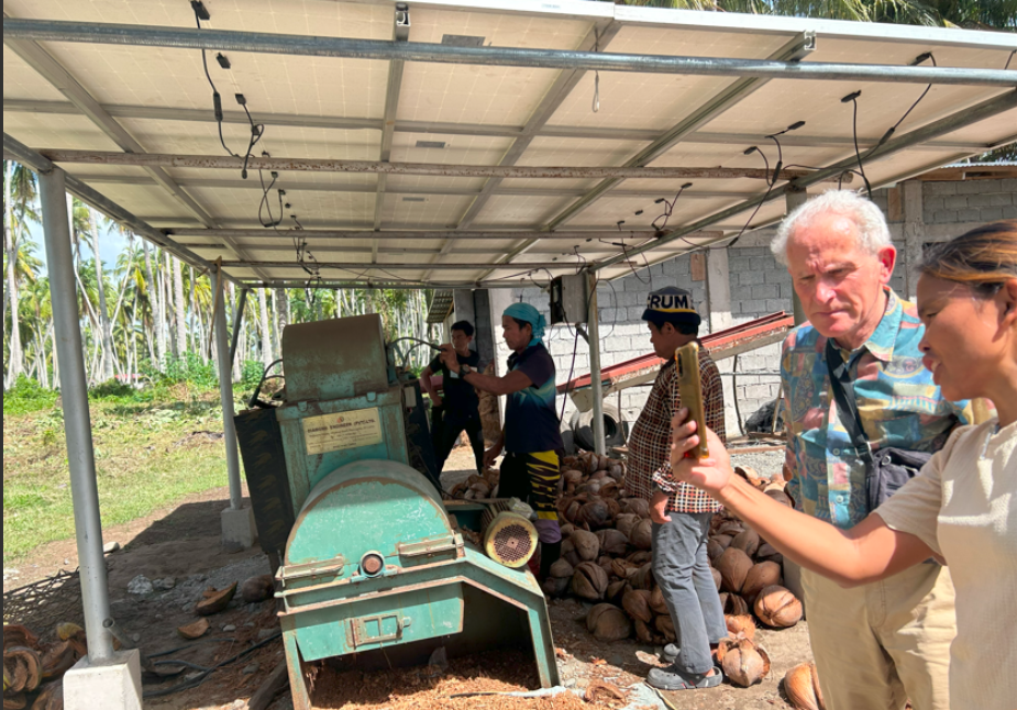 Workers operate machinery while discussing with PUM expert Mr. Cees de Kreij, surrounded by coconuts in an outdoor setting.