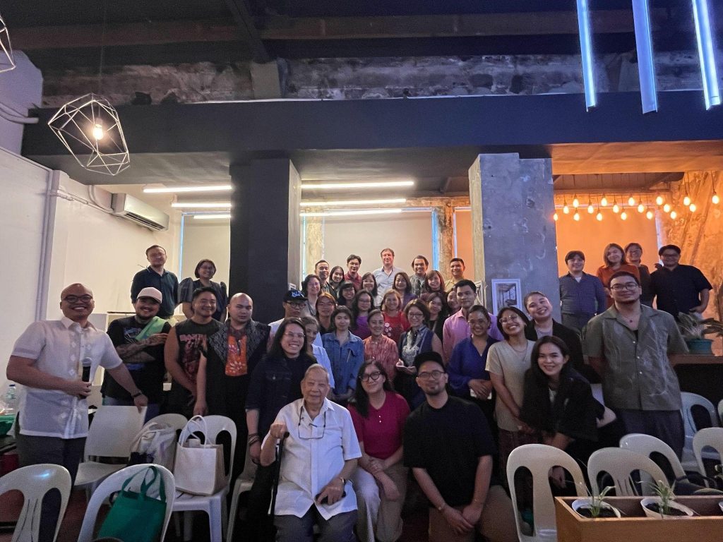 A group of people posing for a photo in a modern industrial room with exposed brick walls and geometric light fixtures. Some individuals are standing while others sit on white plastic chairs. The event, hosted by the DTI's Creative Industries Development Office in partnership with the British Council Philippines, is part of the ACE Hybrid Creative Economy Policy Training Programme held on 12 February 2025 at the First United Building, Escolta Street, Manila. Photo courtesy of Ms. Sari Molintas from the British Council.