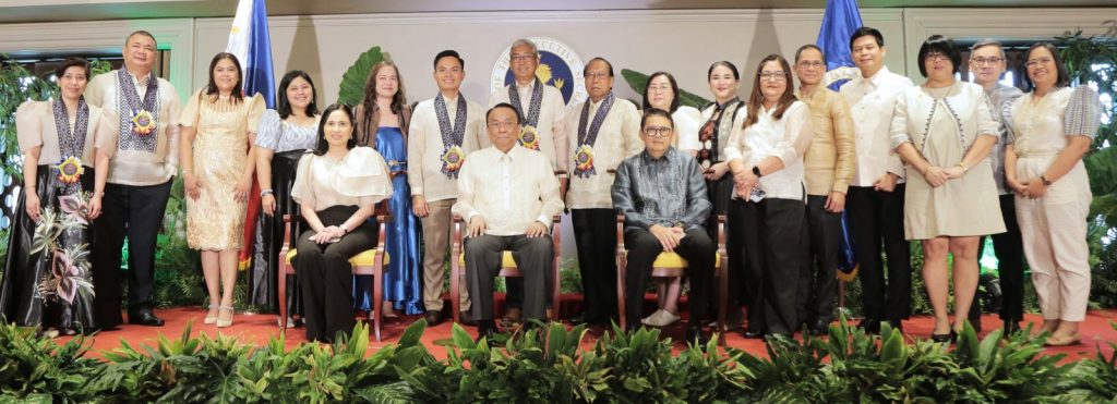 Awardees and officials pose for a formal photo at the 2025 Presidential Awards ceremony in Malacañang Palace, featuring Executive Secretary Lucas P. Bersamin and DTI Secretary Cristina A. Roque, with honorees wearing traditional attire and medallions under the Philippine flag and official seal