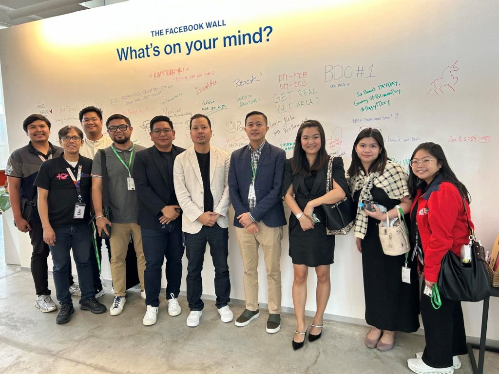 Group of DTI and Meta representatives pose in front of a wall with handwritten notes. The wall reads "The Facebook Wall, What's on your mind?".