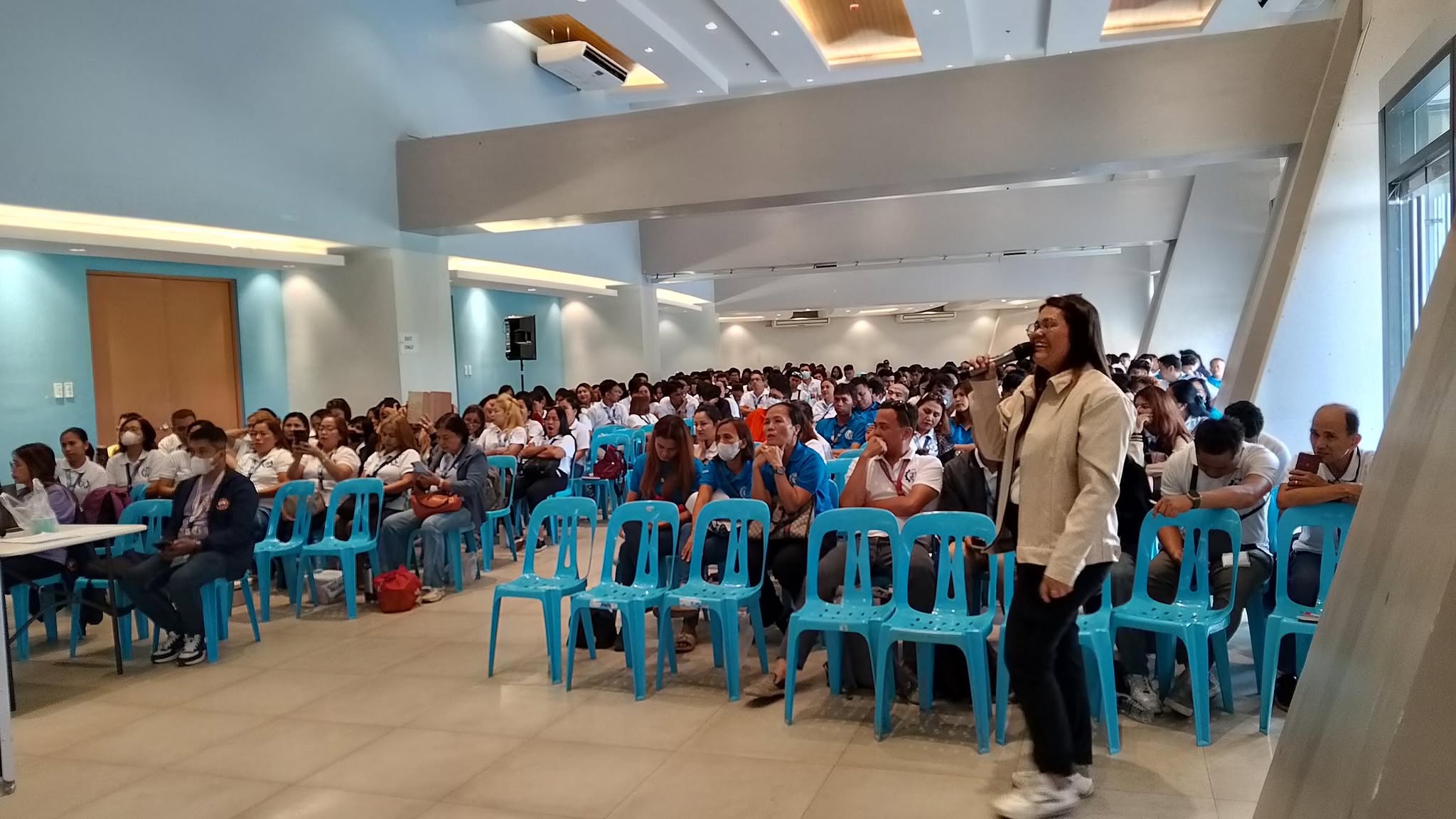 A speaker is addressing a large group of attendees at an indoor event, possibly a seminar, workshop, or conference. The setting suggests a formal or educational gathering.