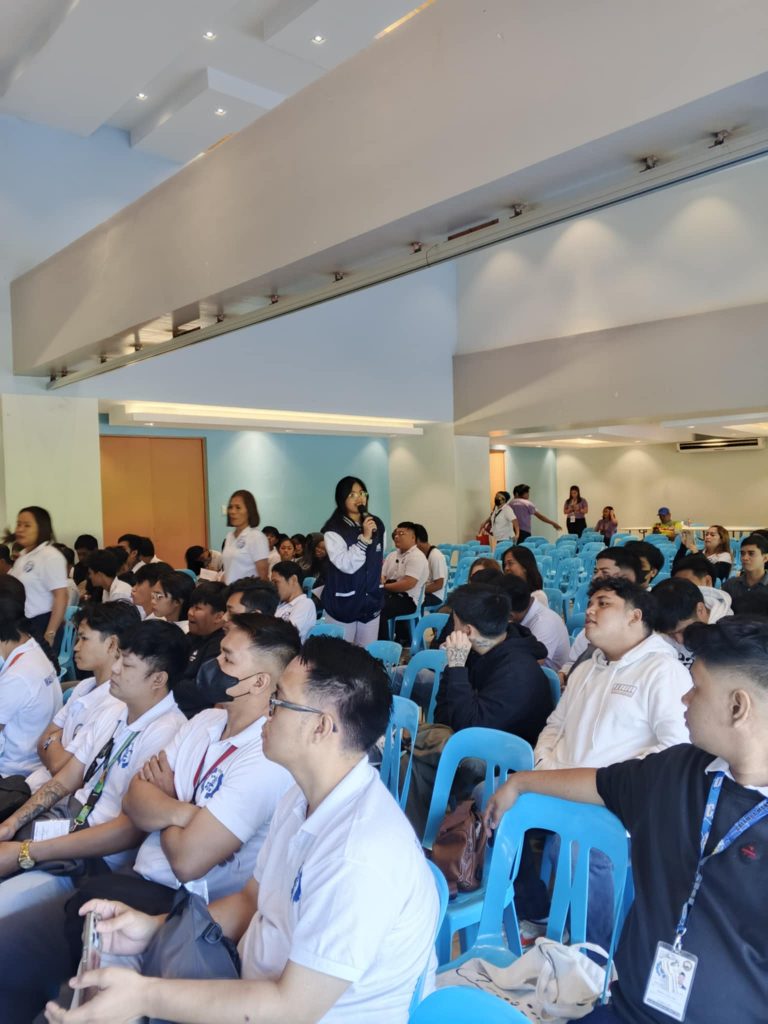 Attendees are participating in an indoor event, possibly a seminar or lecture. A speaker is addressing the group, and participants appear to be engaged in listening or taking notes.