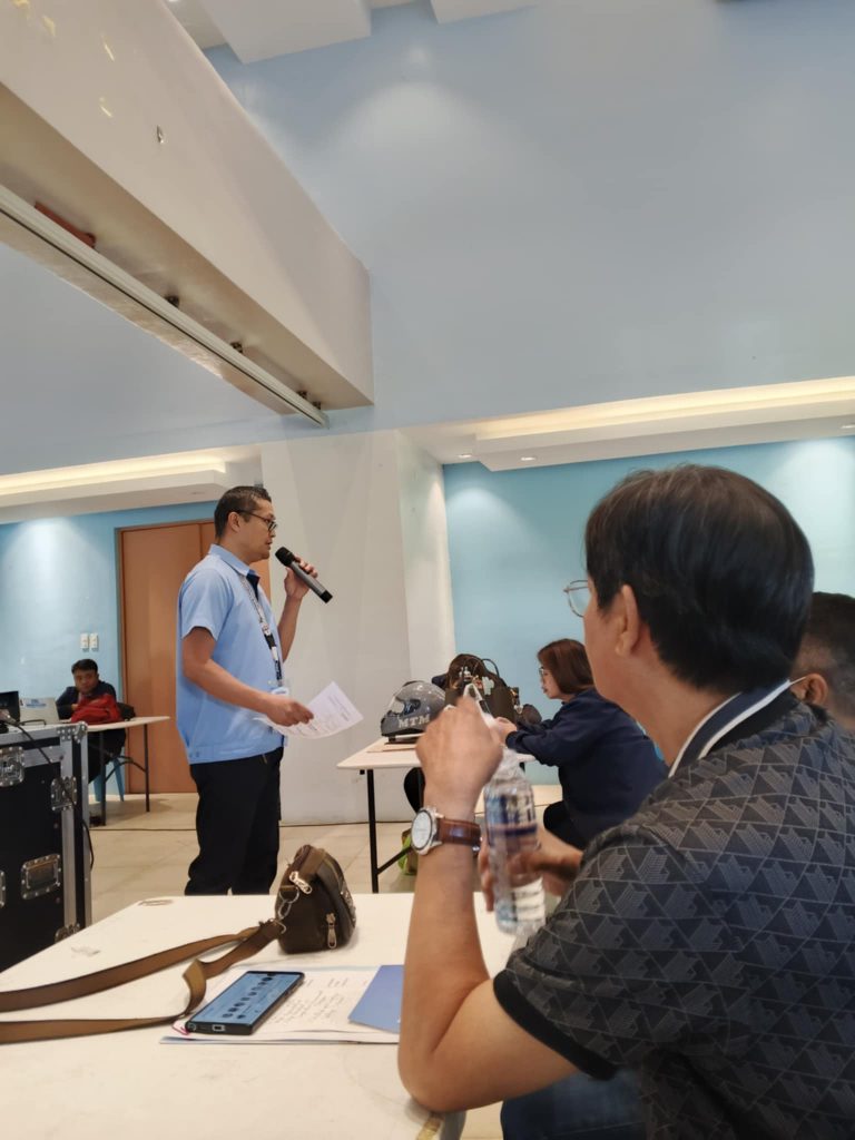 A man wearing a light blue polo shirt and glasses speaks into a microphone, holding papers, at the front of a room with white tables. Other people are seated at the tables, listening. A black equipment case is visible in the foreground.