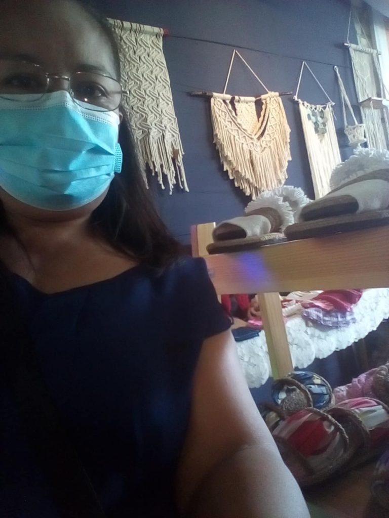 A selfie-style shot shows a person wearing glasses and a light blue surgical mask, partially obscuring their face. They appear to be indoors, possibly in a shop or craft store, given the background. Behind them, various handmade macrame wall hangings in off-white or beige tones are displayed on a dark blue wall. To the right, a wooden shelf holds what seem to be more handcrafted items, including slippers with fluffy white tops. Below the shelf, other woven or textile products are visible. The overall impression is that the person is likely browsing or working in a store that sells artisanal, possibly Filipino-made, goods, considering the likely location.