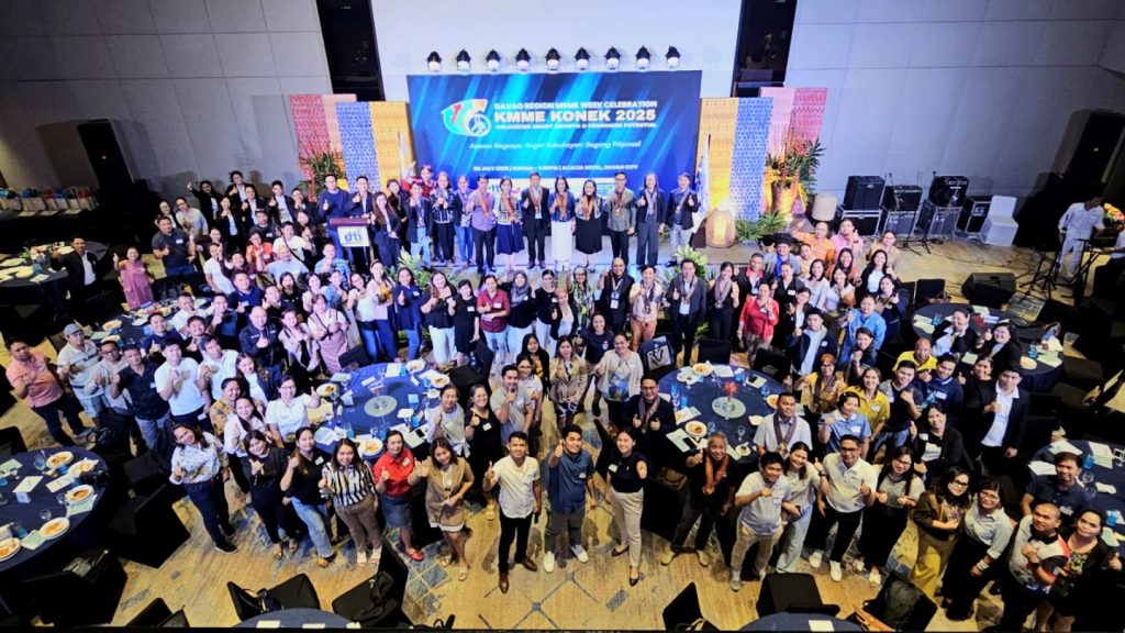 A large group of people gathered in a spacious event hall for the Davao Region's 1st regional mentorship event for MSMEs. The attendees are posing for a group photo, with some standing on stage and others around round tables. A banner in the background reads "KMME KONEK 2023".