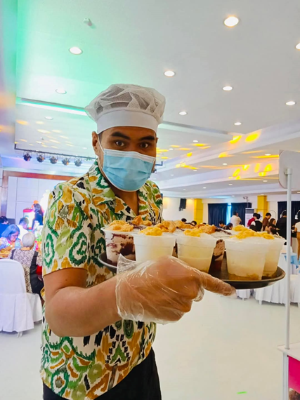 A server in a patterned shirt, chef's hat, face mask, and gloves holds a black tray filled with several transparent plastic cups of halo-halo, a traditional Filipino dessert with layers of shaved ice, sweet toppings, and leche flan. 