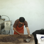 Man in orange apron sorts cocoa beans on a wooden table next to chocolate processing equipment.
