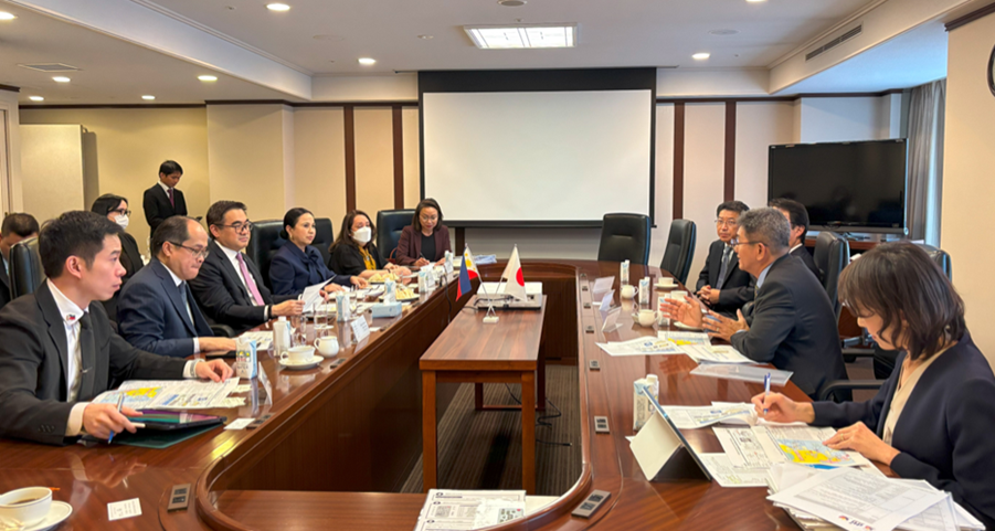 A meeting between Philippine and Japanese officials is taking place around a conference table. Attendees include DTI Assistant Secretary May Niña Celynne Layug, STR Dita Angara-Mathay, Secretary Cristina A. Roque, SAPIEA Frederick D. Go, Director General Tereso Panga, Assistant Secretary Englebert Josef Chua, Mr. Joselito B. De Torres, Executive Officer and Division Manager, Ibiden Philippines Inc., Mr. Naohiko Yajima, Executive Senior Management, Vice President of Electronics Operation, Ibiden Co., Ltd., and Mr. Yuzo Yamagami, President and CEO, Ibiden Philippines Inc. The flags of Philippines and Japan are on the center table. Participants review documents and engage in discussion.