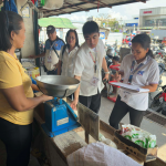 At a busy market in SOCCSKSARGEN, Philippines, two officials from the Department of Trade and Industry (DTI) monitor rice prices. One official in a white uniform shirt and ID badge holds a clipboard looking at it, while the other official in a white shirt stands nearby, observing a vendor in a yellow shirt. who is using an analog scale to weigh rice.