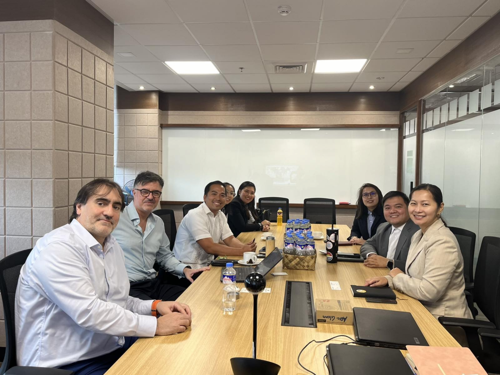 A group of eight individuals seated around a conference table in a modern meeting room. Laptops, water bottles, and a snack basket are on the table. They are wearing business attire and smiling at the camera, appearing to be in a discussion or meeting.