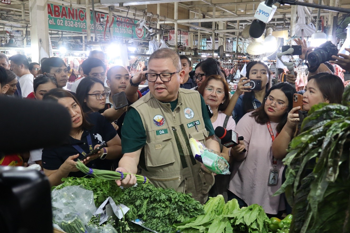 DA Secretary Francisco Tiu Laurel Jr. examines vegetables at a busy market while being surrounded by onlookers and cameras.