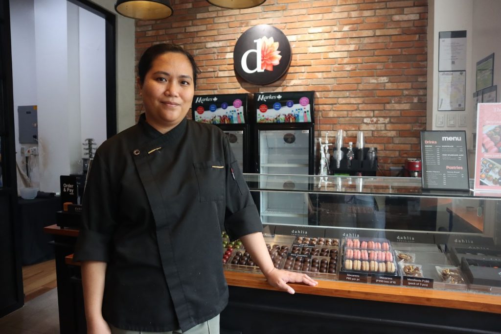 Ms. Aquino of Dahlia, a Filipina chocolatier, stands proudly in her Cebu shop, showcasing chocolate creations and pastries.