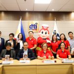 A group of Department of Trade and Industry (DTI) and Jollibee Foods Corporation representatives are in a conference room, holding Jollibee Funko Pop figures. They are posing for a photo during a partnership event with the Philippine flag and the DTI logo in the background, and Jollibee mascot standing with them.